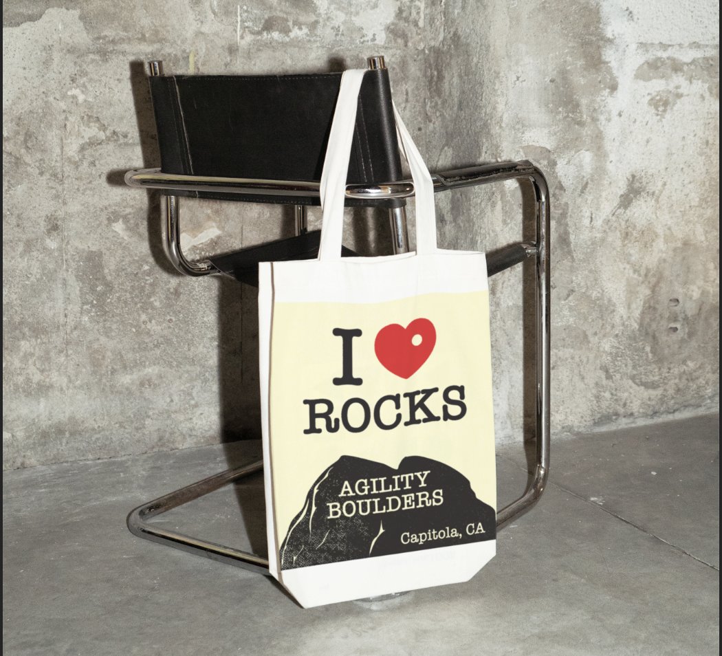 The 'I heart Rocks' design printed on a natural canvas tote bag, hanging from the arm of a black leather and chrome chair against a concrete wall.