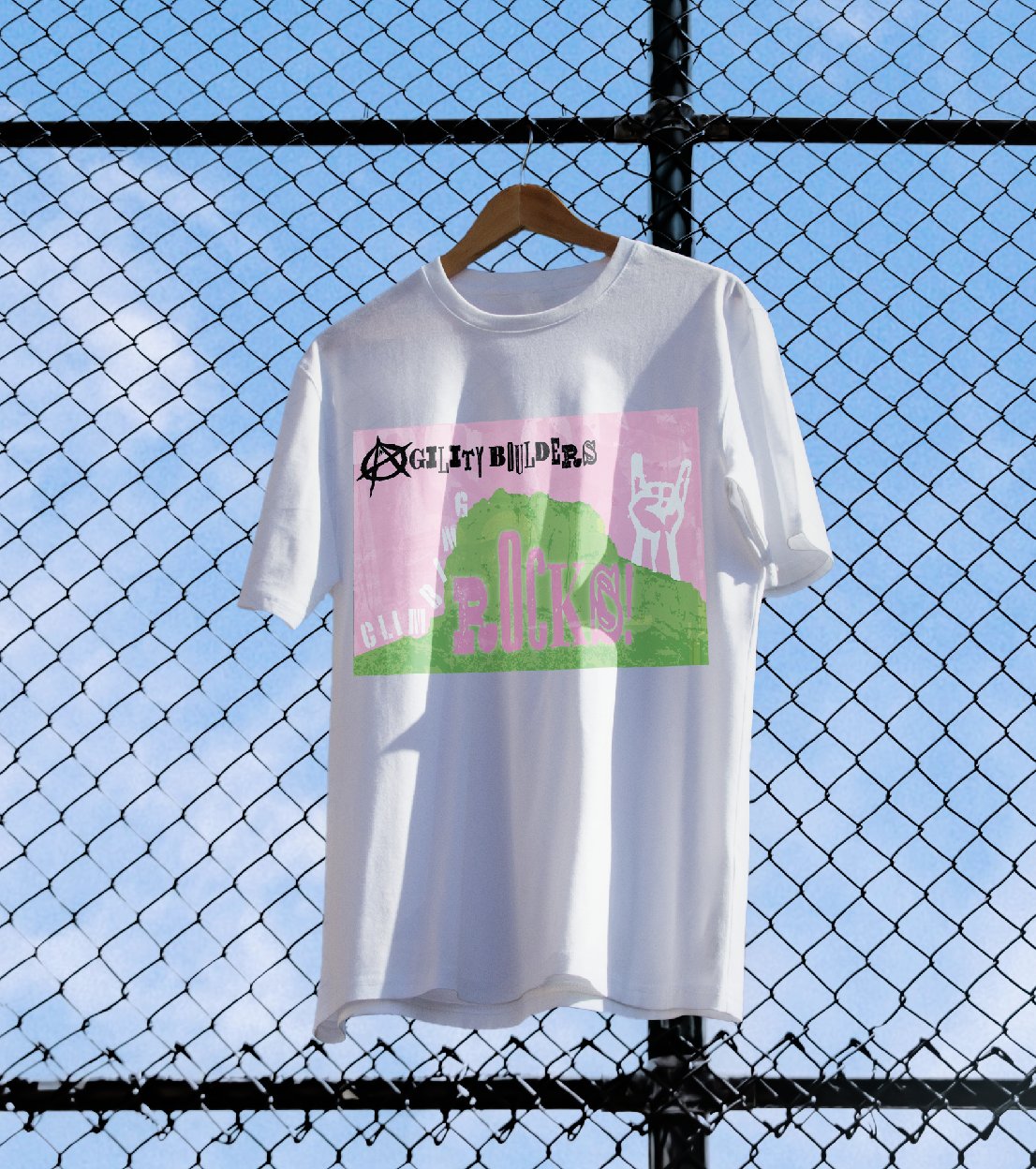 A white t-shirt on a hanger clipped to a chain-link fence against a blue sky, printed with a green and pink boulder graphic.