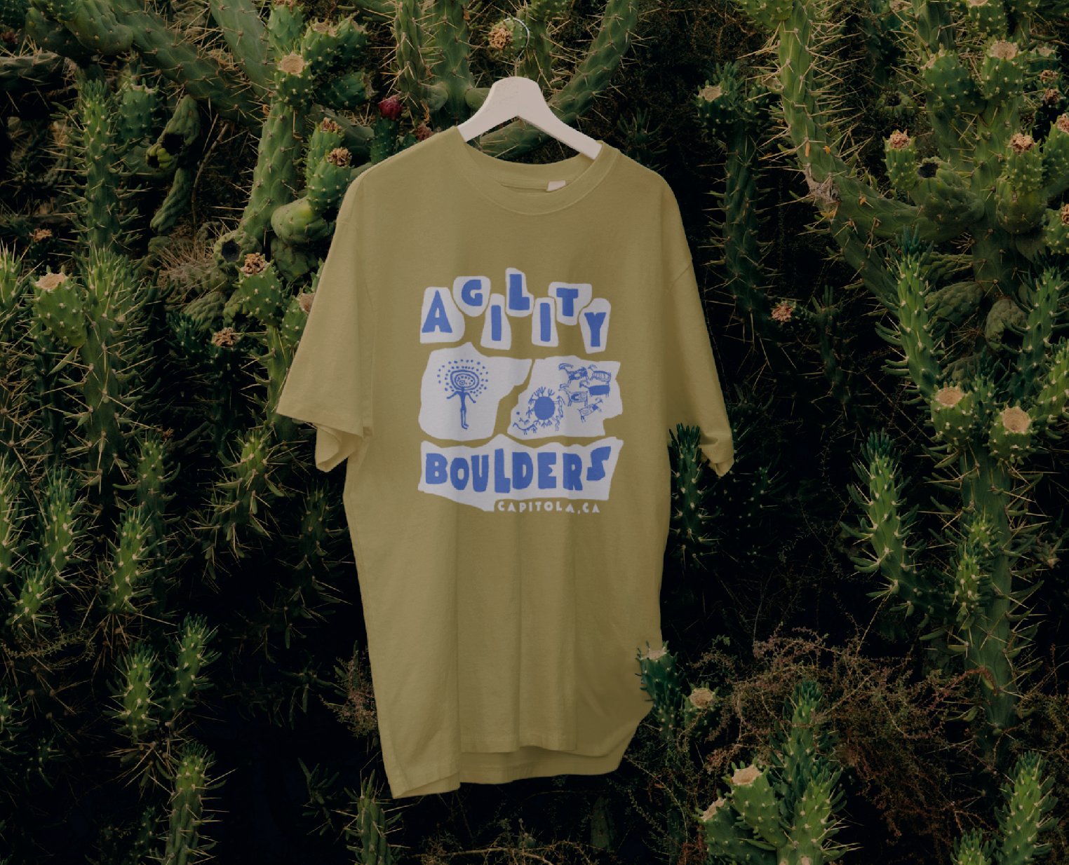 An olive t-shirt hanging in dense green foliage, printed with the blue petroglyph Agility Boulders graphic.