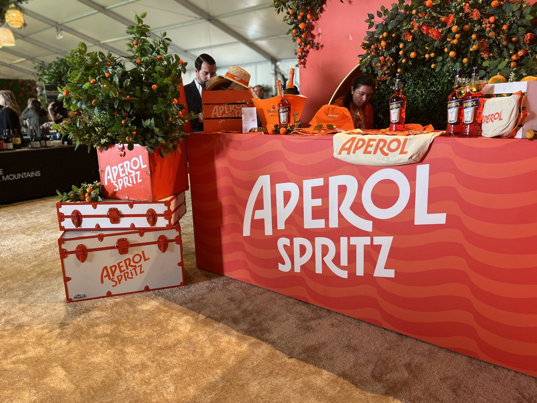 The Aperol Spritz bar under a white event tent: a coral bar front printed with wavy two-tone orange stripes and the Aperol Spritz wordmark, topped with live kumquat trees in coral crates, with white and orange steamer trunks stacked in front.