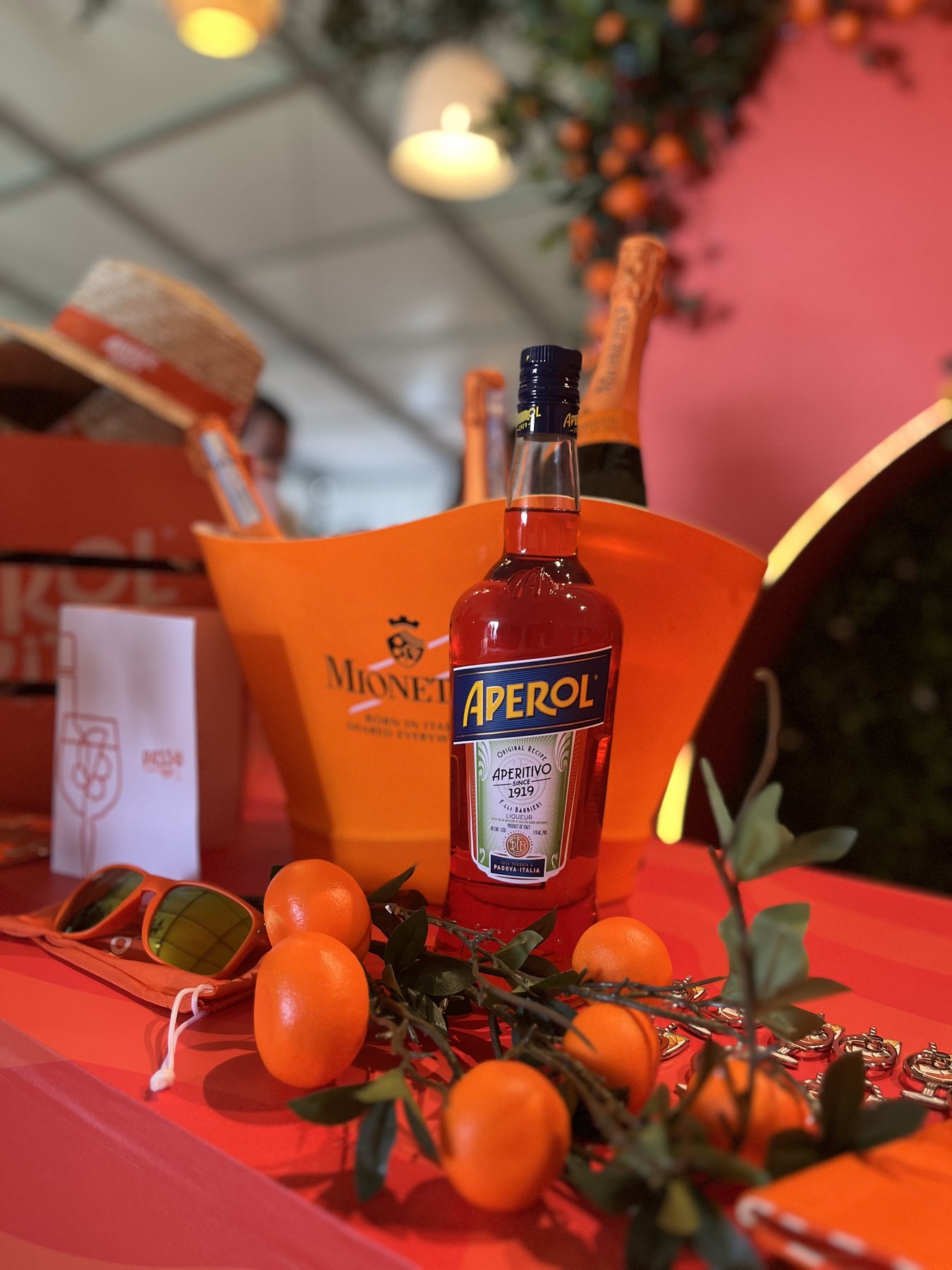 A coral table styled with a bottle of Aperol and prosecco in an orange ice bucket, loose kumquats and cut greenery, orange-tinted sunglasses and a straw boater hat with an orange band behind.