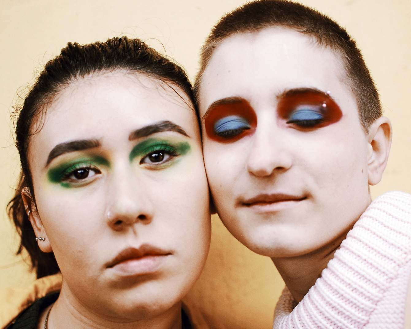 Two faces pressed cheek to cheek against a warm wall, one with green eyeshadow, the other with deep red painted around closed eyes.