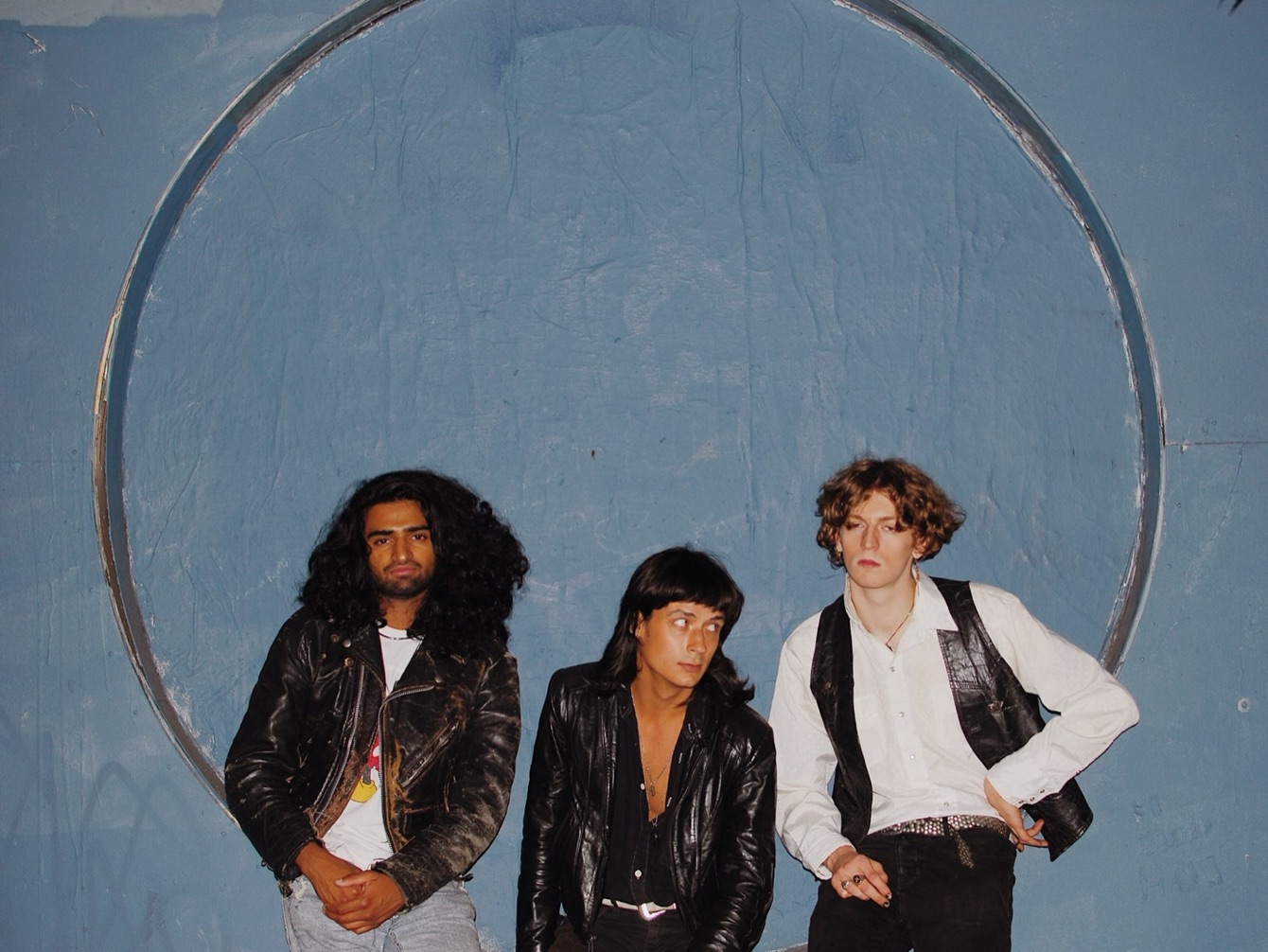 The three members of the Seattle band Karoshi, in leather jackets and a white shirt, leaning against a pale blue wall inside a painted circle.