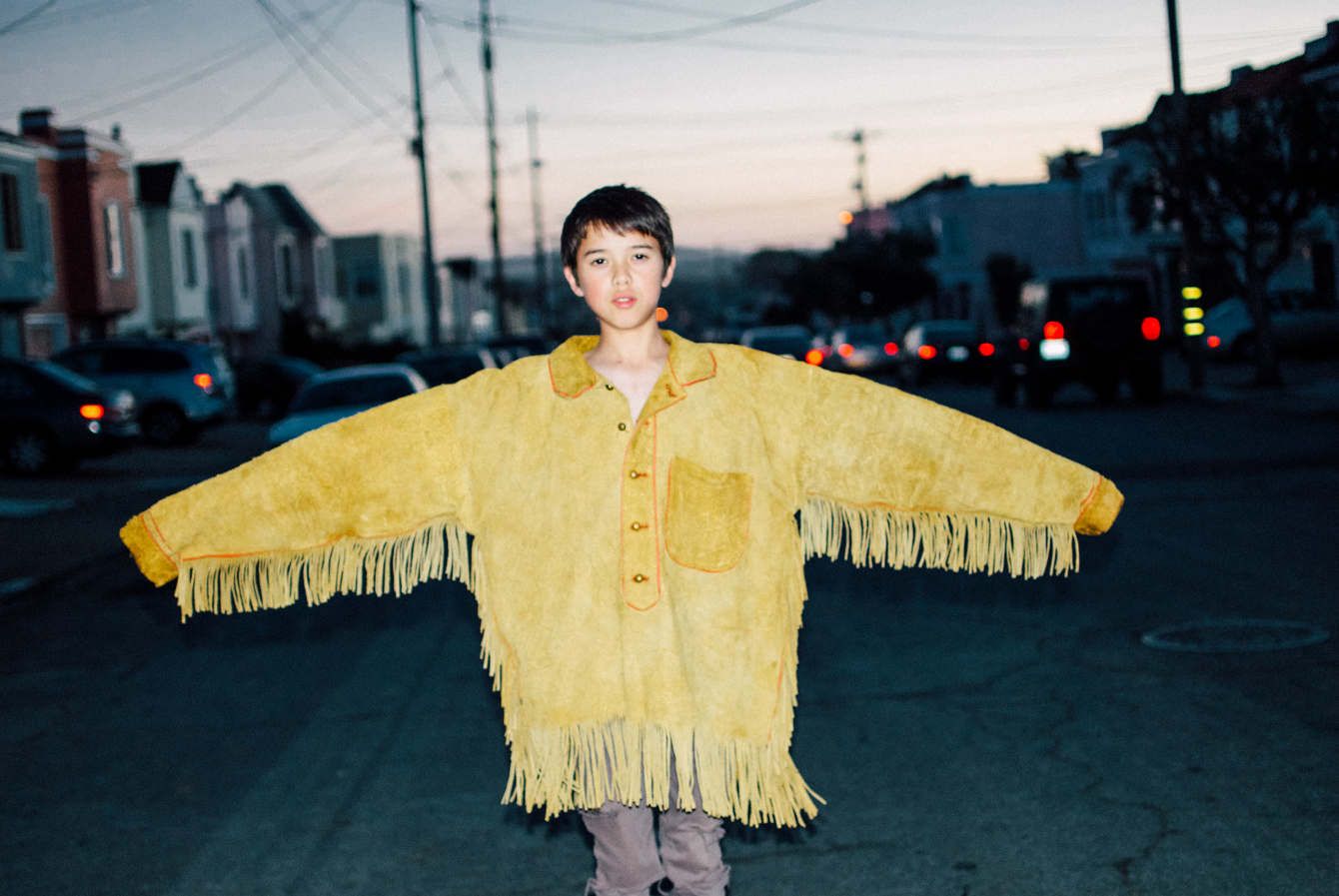 A boy in an oversized yellow fringed suede jacket, arms held wide, standing in a street at dusk under power lines.