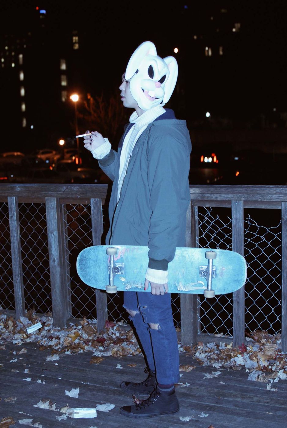A figure in a white rabbit mask and a blue jacket holding a skateboard beside a fence at night, fallen leaves at their feet.