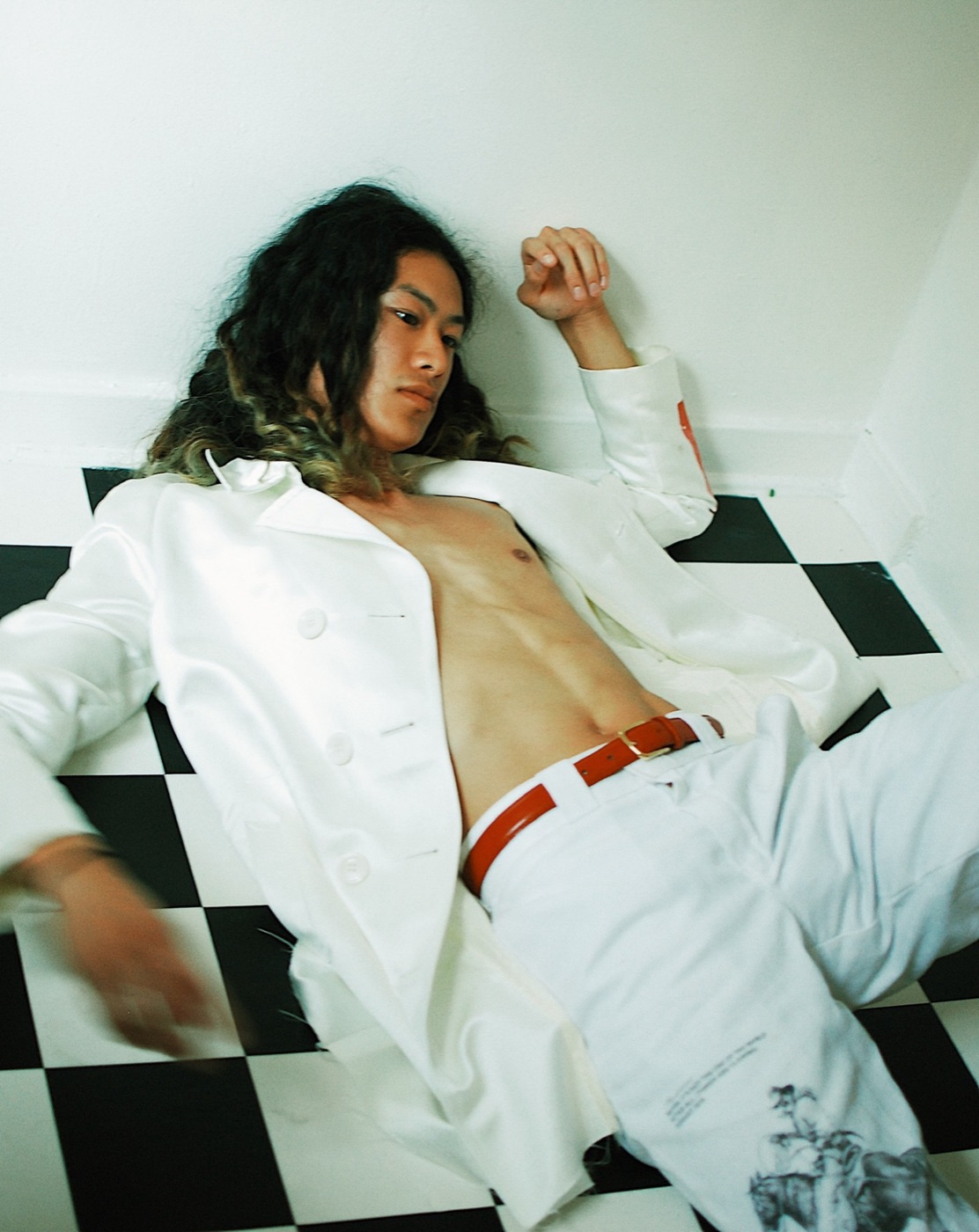A man with long dark hair lying back on a black and white checkered floor in an open white suit and a red belt.