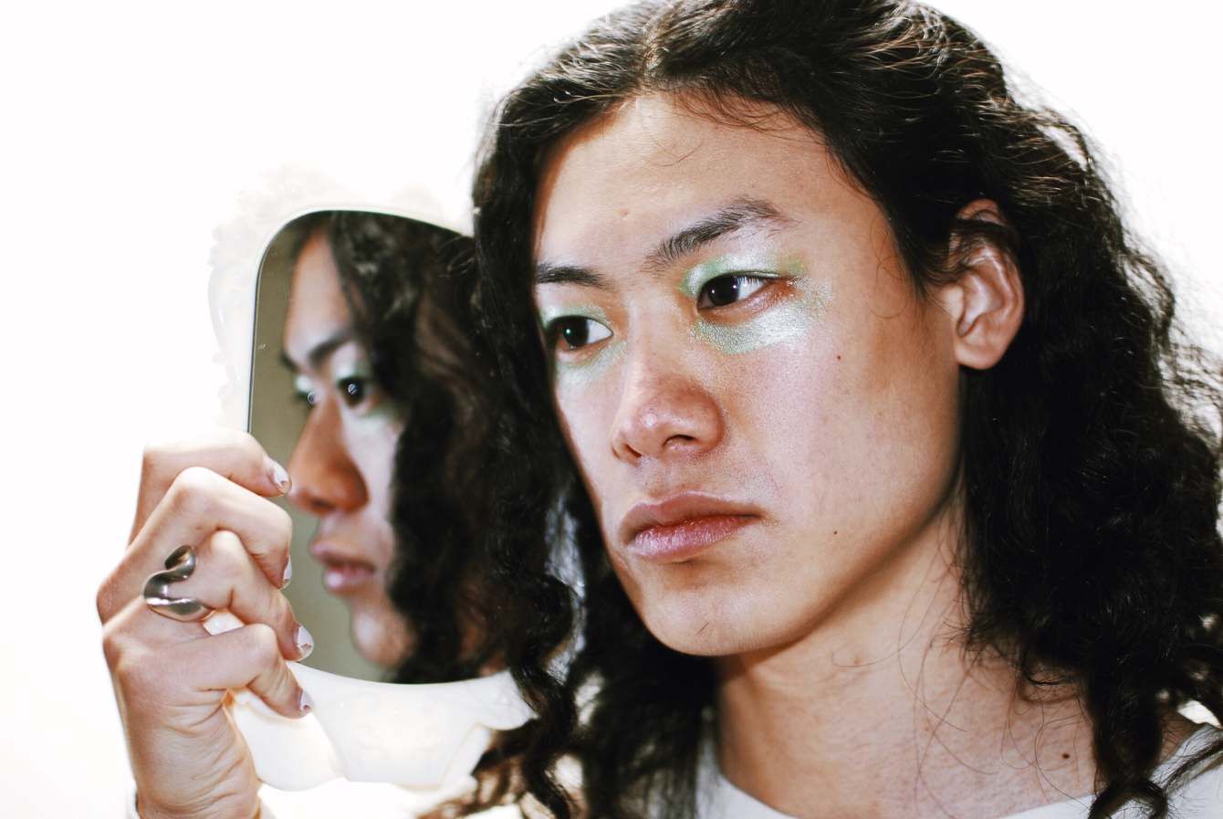 A face with shimmering eyeshadow beside its own reflection in a hand mirror, on white.