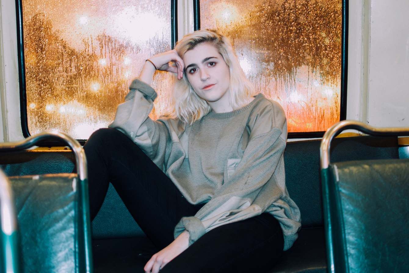 A woman with platinum hair sitting on a bus seat, the rain-streaked window behind her glowing gold.
