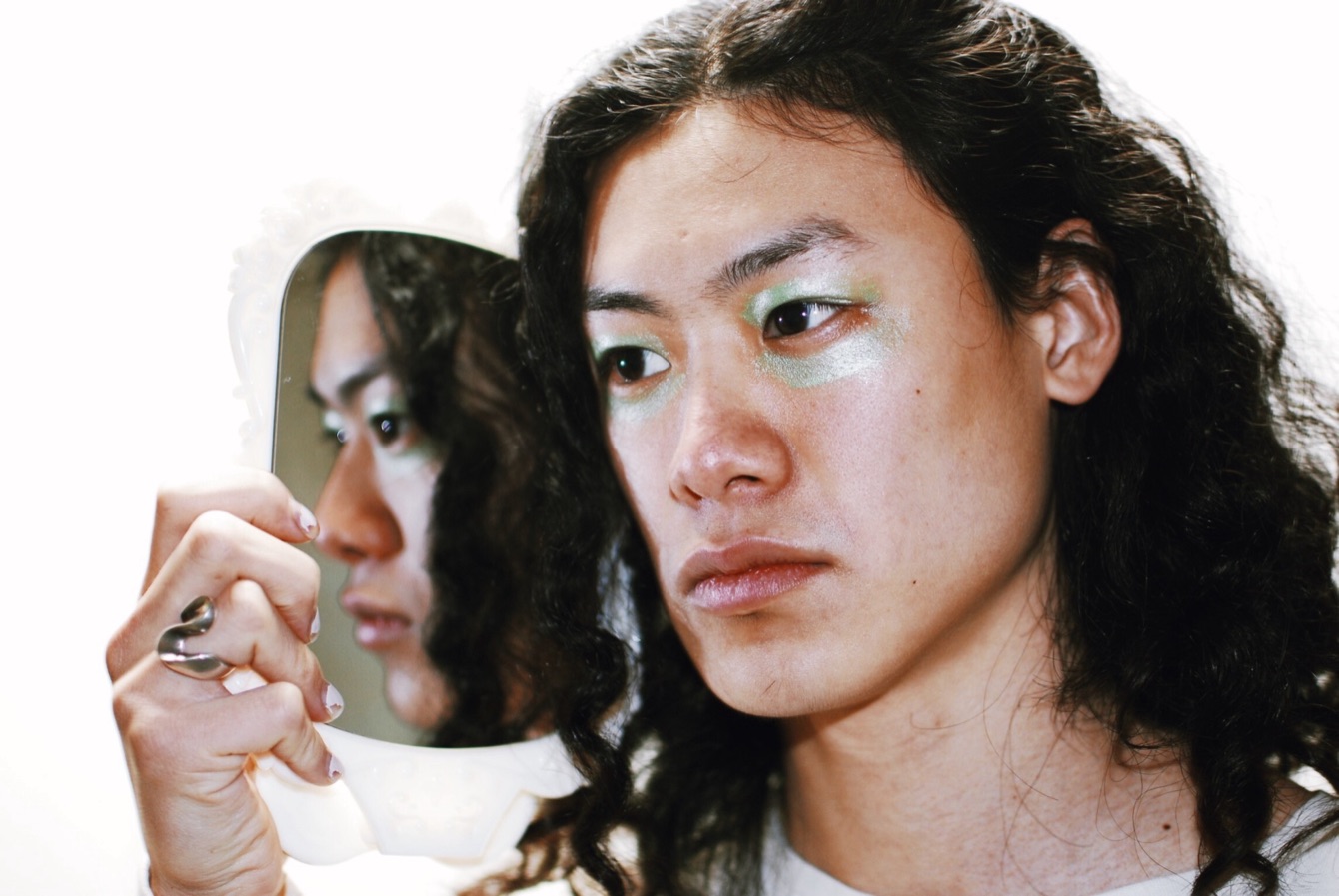 A face with shimmering green eyeshadow beside its own reflection in a hand mirror, on white.