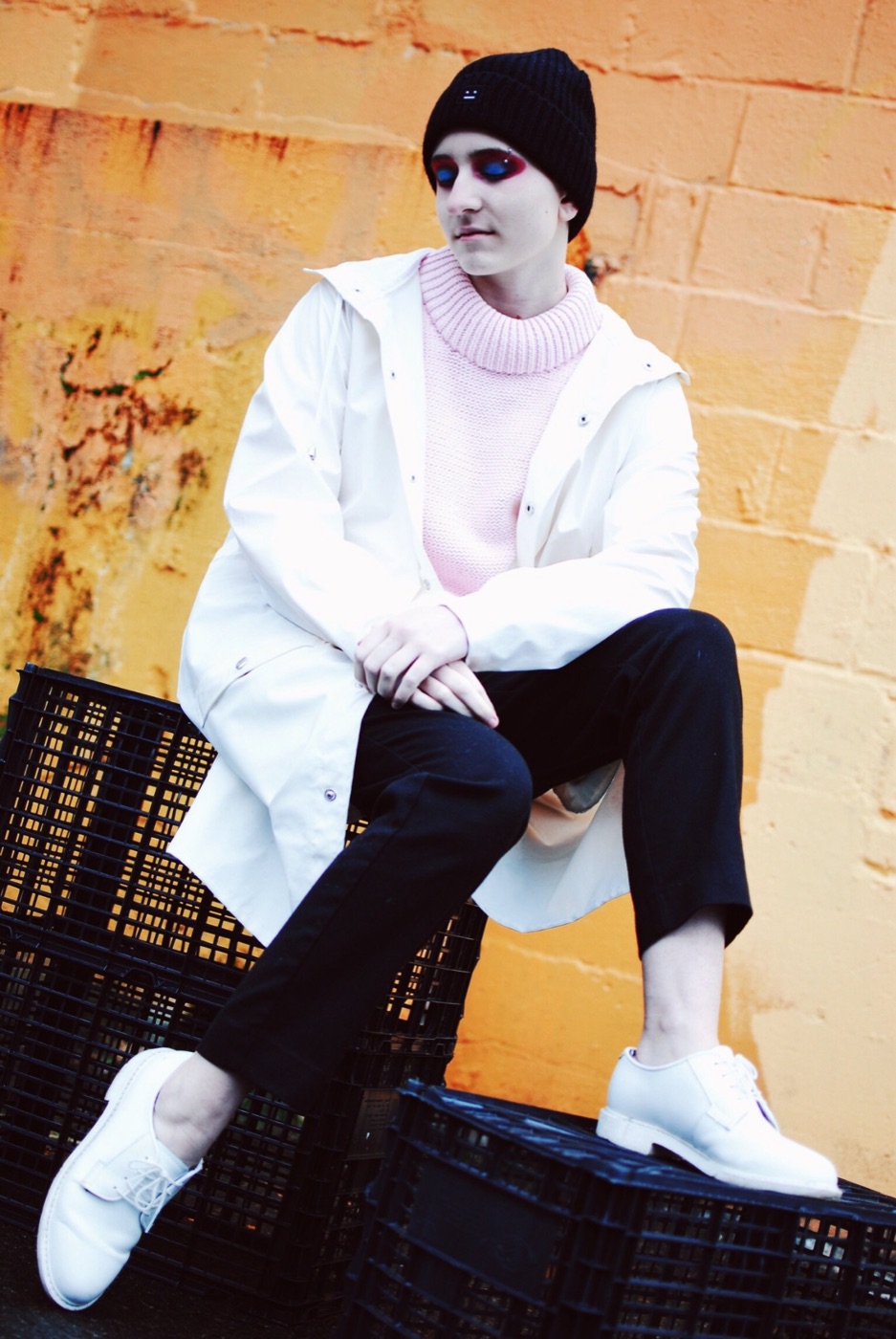 A model in a white raincoat, pink knit and black beanie, deep red painted around closed eyes, sitting on black crates against an orange wall.