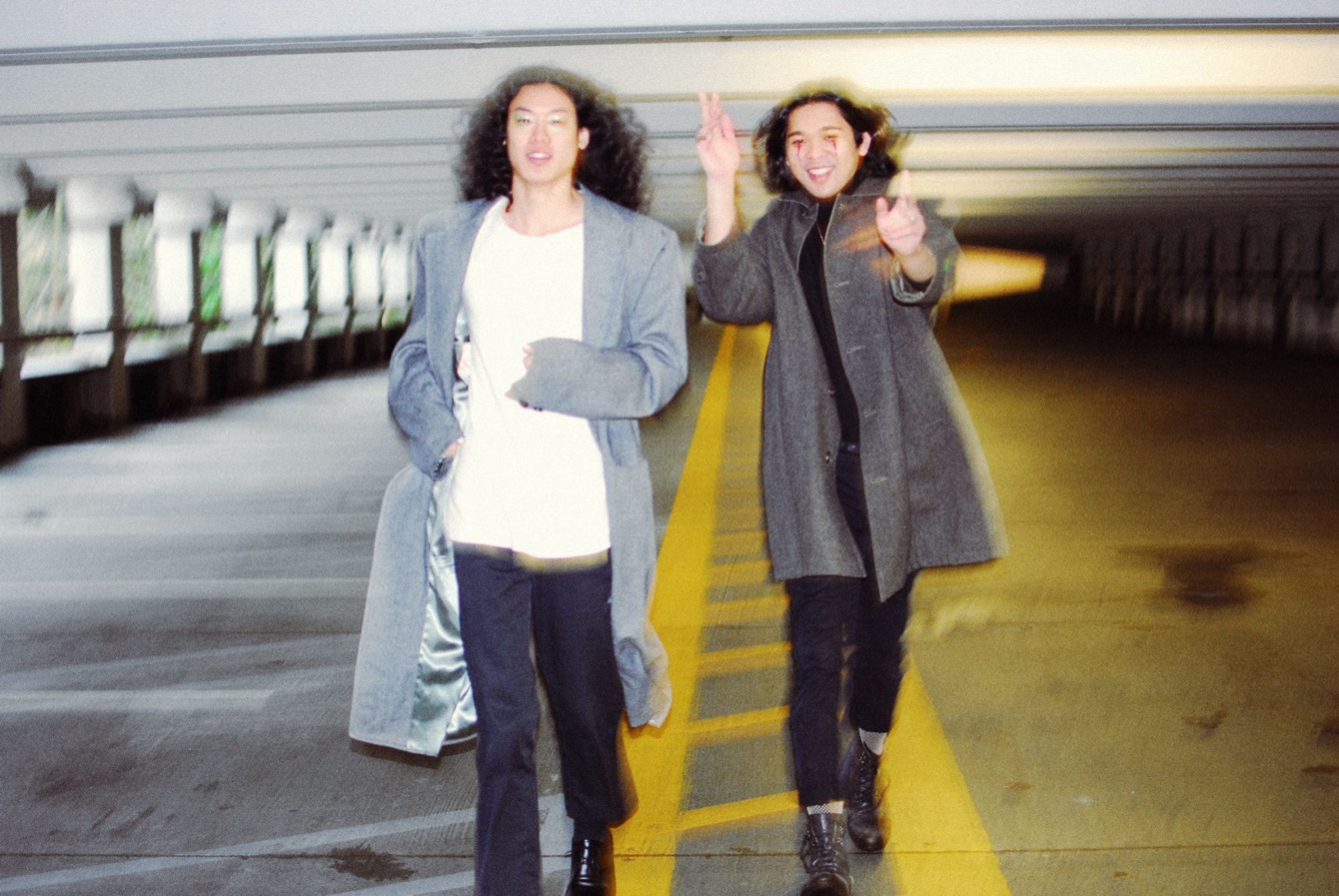 Two friends in long grey coats walking toward the camera along a yellow line in a parking garage, one waving.