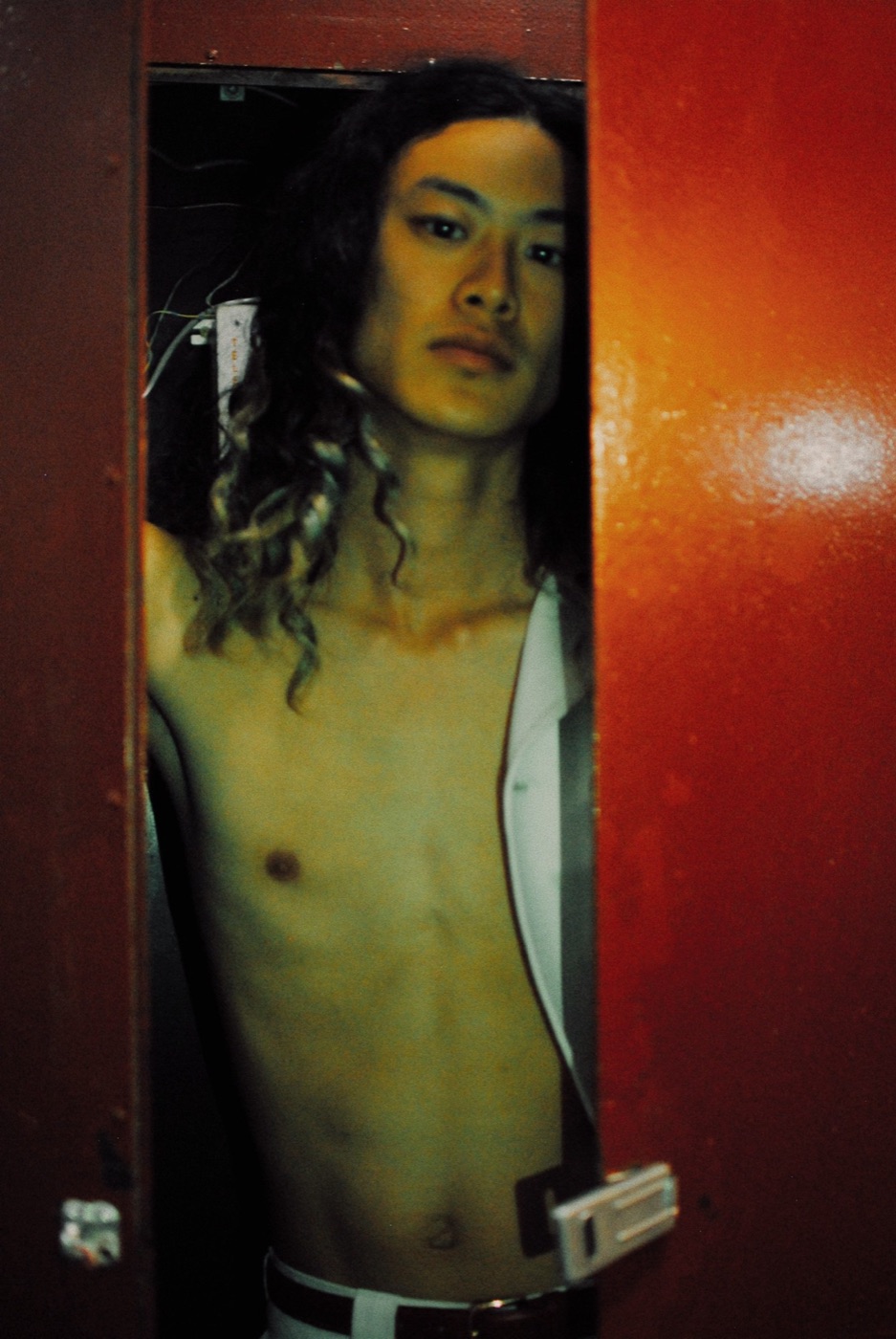 A man with long curling hair standing shirtless in the narrow opening of a red door.
