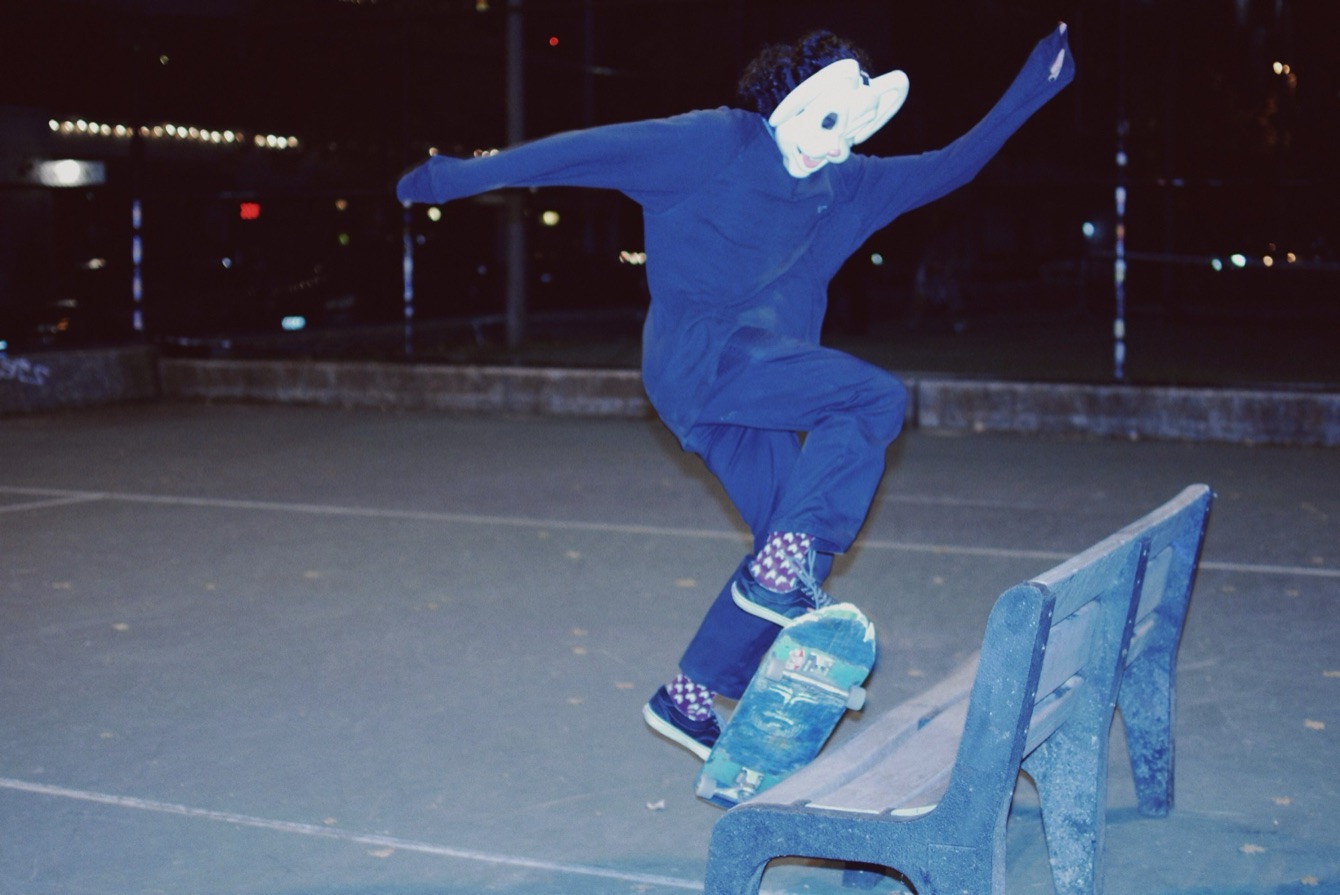 A skater in a white rabbit mask and a blue jumpsuit jumping onto a park bench at night.