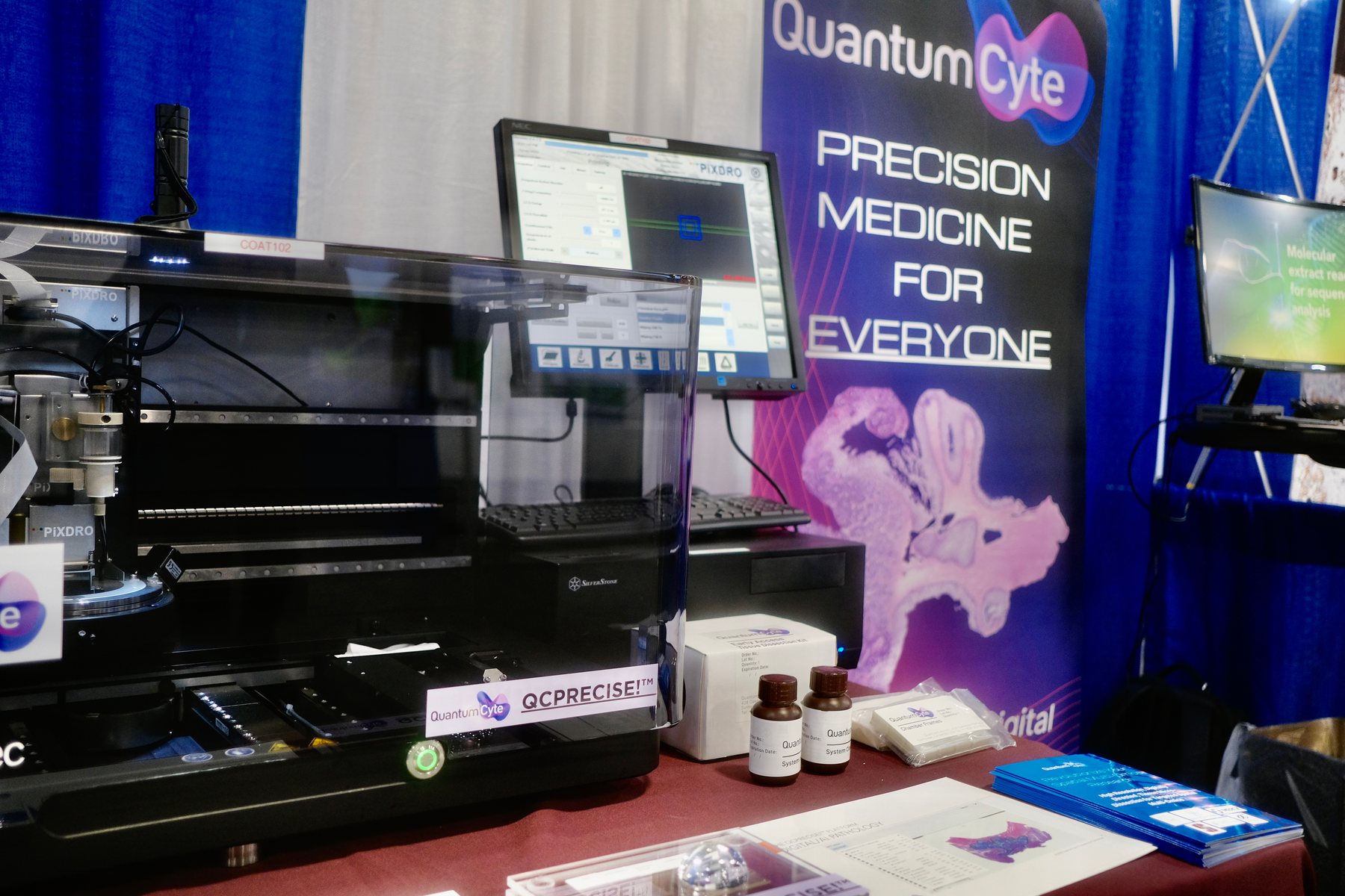 Close view of the QuantumCyte booth: an automated microdissection instrument beside the pull-up banner reading Precision Medicine for Everyone.