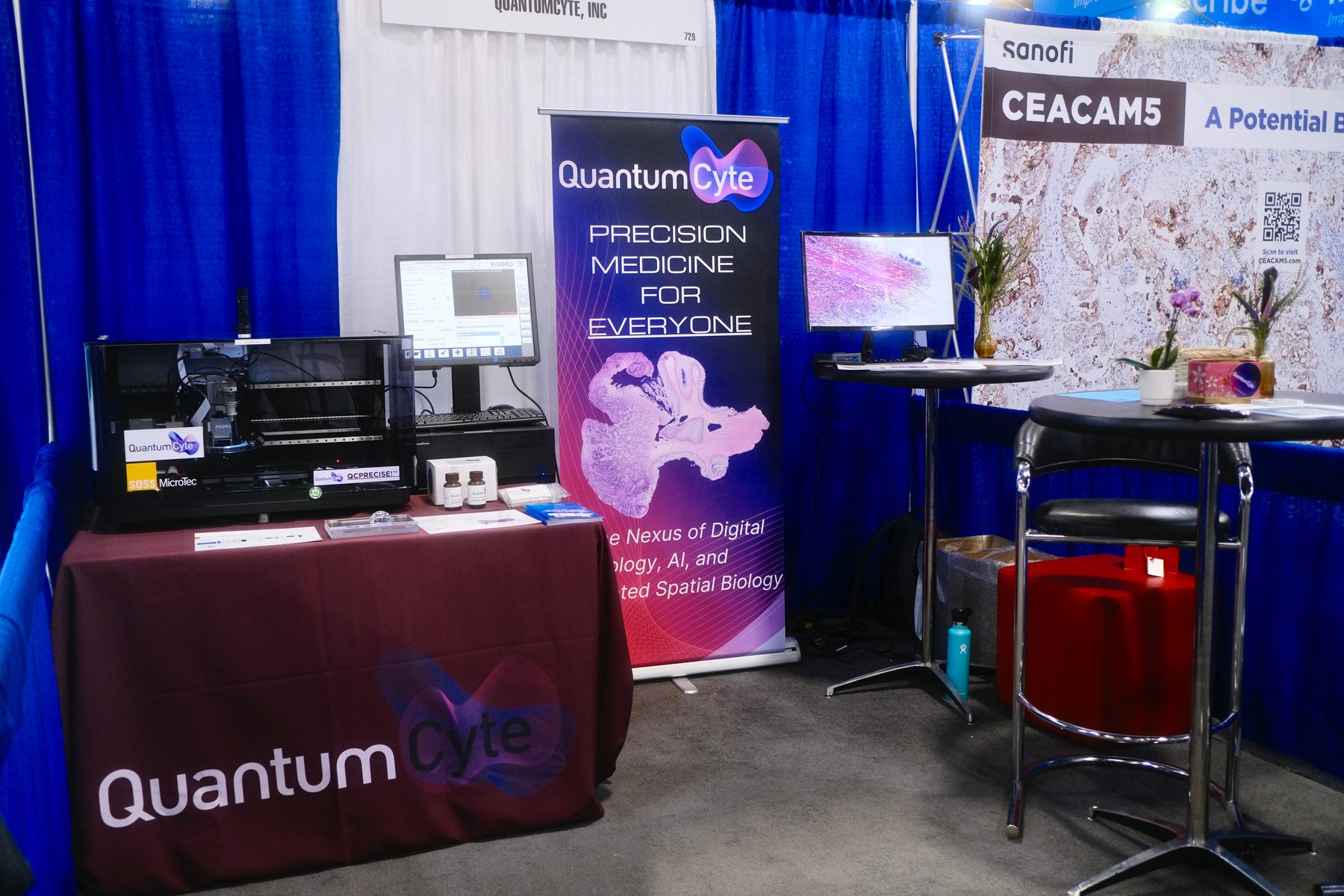 The finished QuantumCyte booth at the tradeshow: a draped table with the logo, a microscope workstation, a monitor, and a pull-up banner reading Precision Medicine for Everyone.