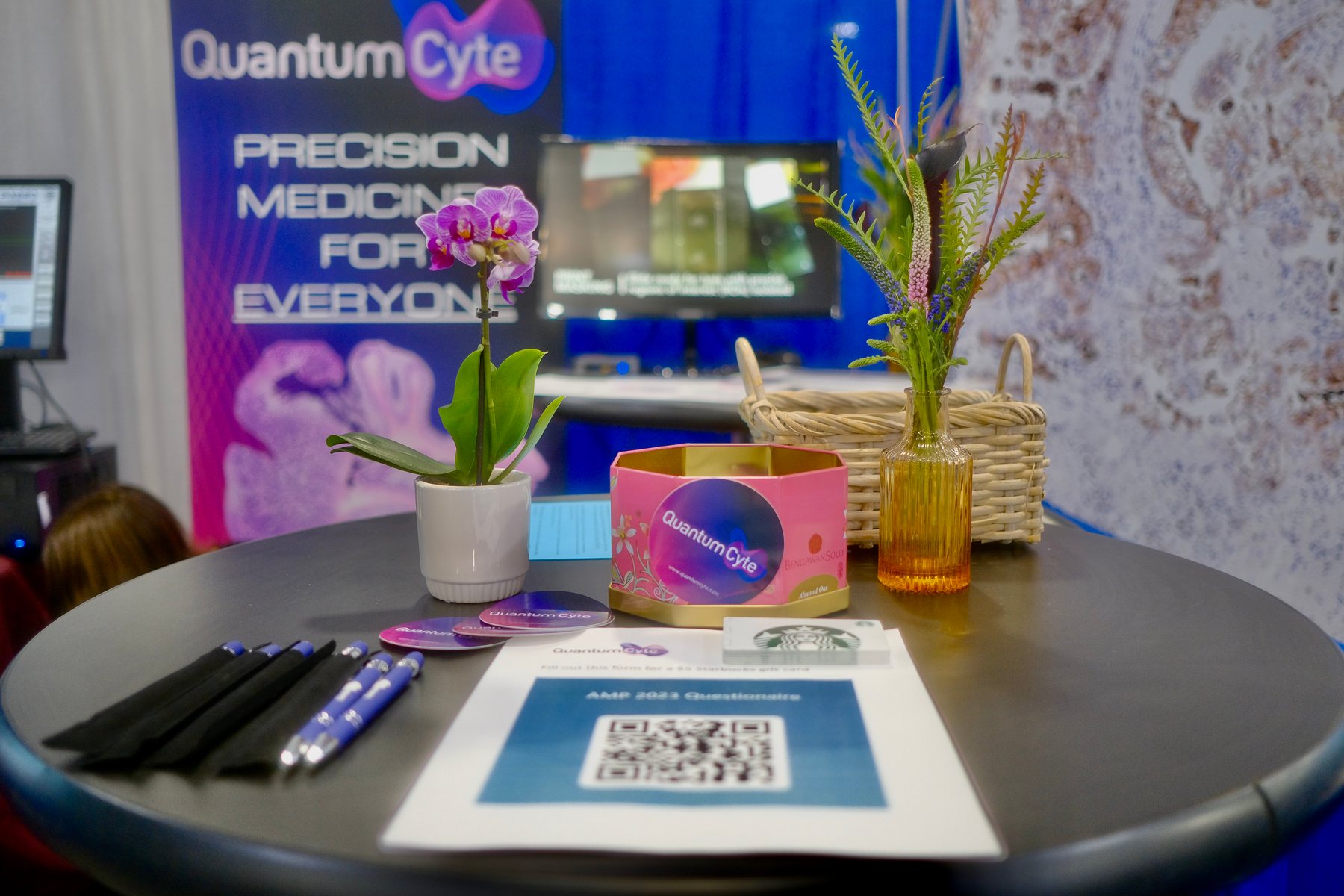 The QuantumCyte cocktail table at the booth, set with orchids, branded pens, a round logo tin and a QR code card, in front of the backdrop.