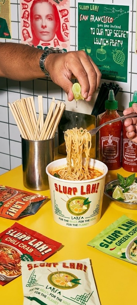 A hand squeezing lime over a Slurp Lah cup of laksa noodles, with chopsticks, chili sauce and Slurp Lah packets on a yellow table.